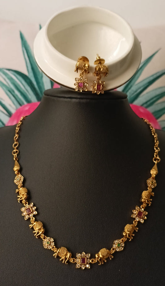 Gold necklace and earrings on a black mannequin with a white bowl and pink flower in the background.