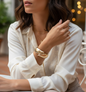 Woman sitting at a table outdoors wearing a white shirt and gold bracelet.